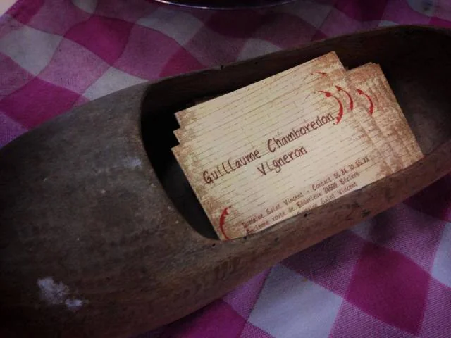 Detail artisanal : cartes de visite en bois 'Guillaume Chamboredon Vigneron' presentees dans un vieux sabot en bois, sur une nappe a carreaux. Touche authentique et conviviale, valorise l'identite artisanale du vigneron (640x480). Source Herault Tourisme.