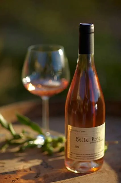 Belle photo lifestyle : bouteille de rose 'Belle Rose' avec un verre rempli pose sur un tonneau, lumiere doree de fin de journee, bokeh vert, brindille d'olivier en avant-plan. Packshot d'ambiance soigne et chaleureux (426x640). Source Herault Tourisme (galerie du domaine, ancien nom Saint-Vincent).
