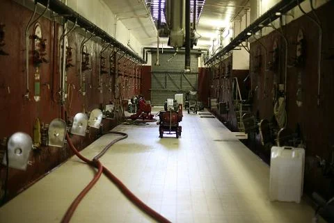 Interieur du chai de la cave cooperative : longue halle industrielle bordee de cuves de fermentation en beton de chaque cote, sol carrele, tuyaux au sol et petit chariot rouge au centre. Vue authentique de l'outil de vinification cooperatif, mais cadrage utilitaire, eclairage faible et basse resolut